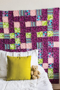 pink quilt hanging behind a white bed with a yellow pillow and a teddy bear